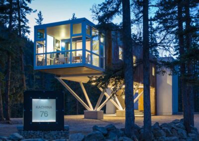 A modern house elevated on stilts is surrounded by tall trees. Large windows on one side reveal the interior, illuminated against the evening sky. A sign at the base reads "Kachina 76." The structure rests on a solid foundation with rock landscaping below.