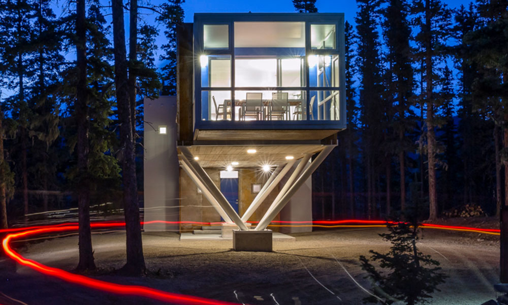 Modern elevated house with large glass windows, nestled among tall trees at night. The house stands on steel supports and has a lit interior. Red and white light trails from a vehicle encircle the structure, highlighting its unique design.