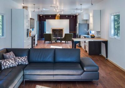 A modern open-concept living and kitchen area featuring a large black sectional sofa in the foreground. The kitchen has white cabinets, stainless steel appliances, and a wooden island. Two green armchairs face a fireplace with a brick surround at the back.