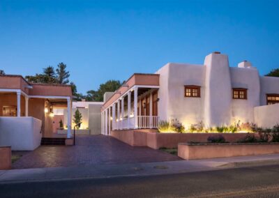 A dusk scene of an adobe-style building with a smooth stucco exterior, wooden accents, and a front porch. The building is illuminated by exterior lights and surrounded by a landscaped area with small plants. The sky is clear and blue.