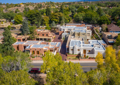 Aerial view of a residential neighborhood featuring houses with flat roofs and surrounded by trees. The buildings have a mix of colors including white, beige, and red. A road runs through the foreground with a red vehicle parked along it.