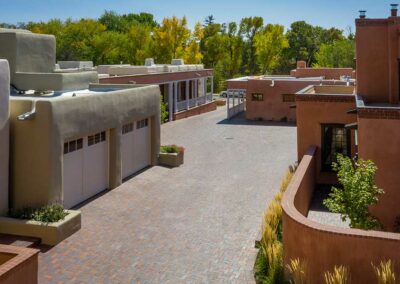 A residential complex featuring Southwestern-style stucco buildings with flat roofs and brick-paved driveways. Trees with autumn foliage are in the background. Several garage doors and small planters with flowers are visible. Clear blue sky above.