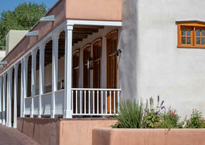 A light-colored building with wooden framed windows and a porch. The porch is supported by white columns and has a white railing. There is a small garden with various plants and flowers in front. The building has a flat roof and a brick pathway beside it.