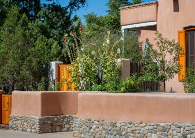 A southwestern-style house with adobe walls and orange shutters is surrounded by tall flowering plants and trees. The front yard has a decorative stone wall and an orange gate. The scene is set under a clear blue sky.