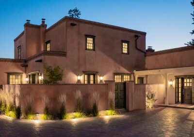 A two-story adobe-style house is shown at dusk with lights illuminating the exterior. The house features flat roofs, small windows, and a courtyard with landscaped plants. The surroundings are calm and serene, with the sky in the background transitioning to night.