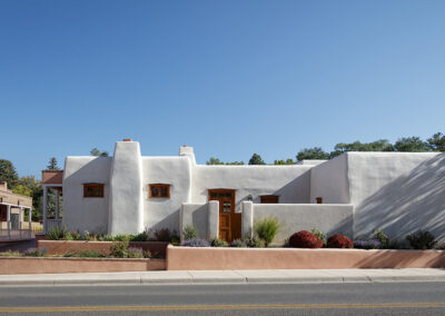 A single-story white adobe building with a flat roof and wooden framed windows stands against a clear blue sky. The structure is surrounded by a low stucco wall, landscaping with shrubs and flowers, and a paved sidewalk with a partial view of a street.