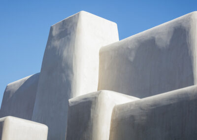 A close-up shot of white stucco buildings with clean, angular lines and smooth surfaces, set against a clear blue sky. The architecture has a minimalist, geometric design with sharp edges and simple forms.