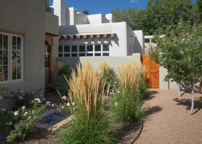 A small garden with tall ornamental grasses and flowering plants is bordered by a gravel path leading to a wooden gate. The garden is adjacent to a building with smooth, light-colored stucco walls, wooden beams, and large windows. Trees are in the background.