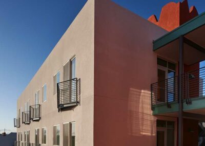 A beige, modern three-story building with a flat roof and metal railings on the balconies is shown against a clear blue sky. Partial views of the structure reveal its minimalist design with clean lines and large rectangular windows.