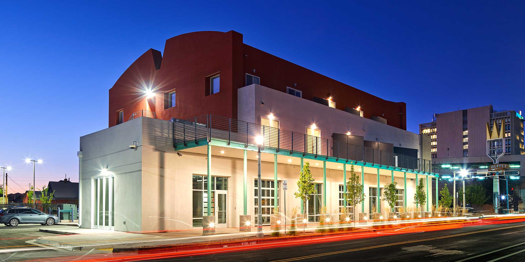 A modern, two-story building with a mix of white and red exterior walls is depicted at dusk. The structure features large windows and a balcony with teal railings. The surrounding area includes streetlights, trees, and a backdrop of additional buildings.