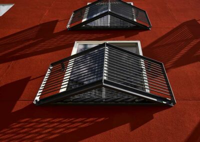 The image shows the exterior of a building with bright red walls and multiple small windows, each framed by an angular, black metal balcony. The shadows of the balconies create geometric patterns on the wall.