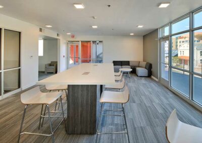 A modern office lounge features a long white table with high chairs around it. The room has large windows, light gray carpet, and a seating area with a sofa and chairs. A glass-walled room and an exit door are visible in the background.