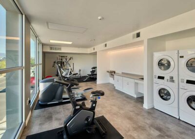 A modern room serving as both a gym and laundry area. There are exercise bikes, weight machines, and large windows. On the right, there are stacked washers and dryers against the wall, with folding counters nearby.