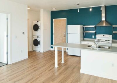 A modern apartment kitchen area with white appliances including a refrigerator, stove, and microwave. There is a washer and dryer stacked in the corner near a door. The room has hardwood flooring, a teal accent wall, and white cabinets with a gray countertop.