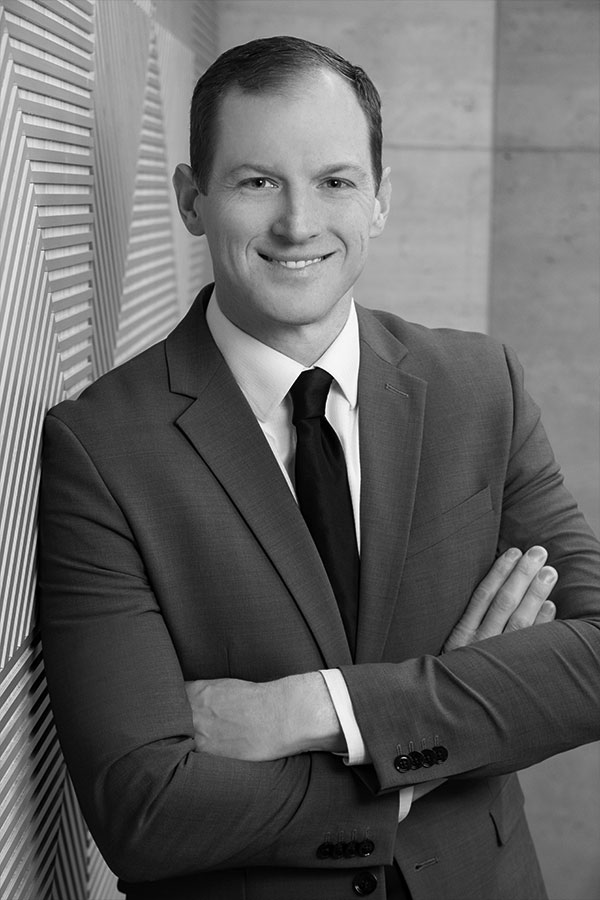 Black and white image of a person wearing a suit and tie. They are standing with arms crossed, smiling at the camera. The background features a textured wall with diagonal lines on one side and a smooth surface on the other.