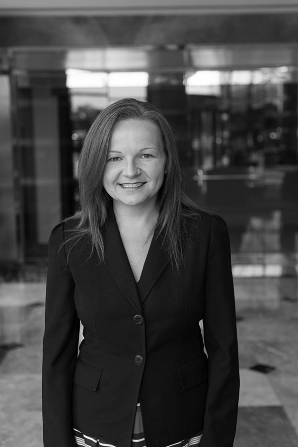 Black and white photo of a woman with long, straight hair smiling at the camera. She is wearing a dark blazer and standing in a polished indoor space with reflective surfaces and a blurred background.