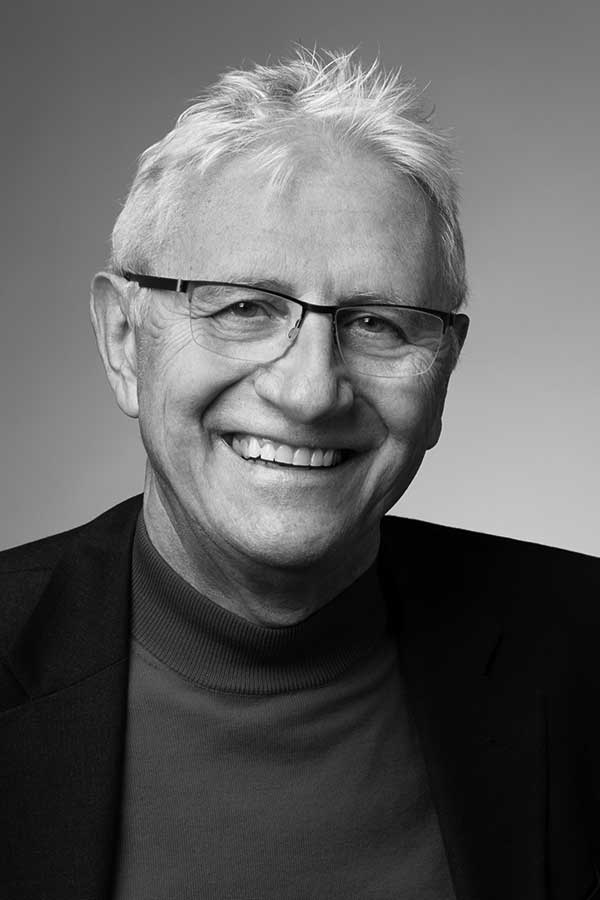 A black and white portrait of an older man with short gray hair, wearing glasses, a dark blazer, and a turtleneck. He is smiling and looking directly at the camera against a plain background.