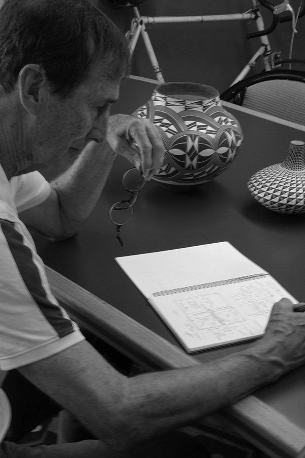 A man is seated at a table, holding glasses in one hand and writing or sketching in a notebook with the other. Two decorative pots and a bicycle are in the background. The image is in black and white.