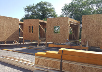 A construction site with partially built wooden structures. Several walls, framed with oriented strand board (OSB), are in place. Plywood and lumber are stacked in the foreground. A person wearing a safety helmet is seated by one of the structures. Trees are visible in the background.