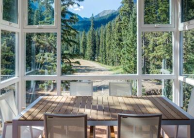 A dining area features a table and chairs surrounded by large windows, providing a clear view of a forest with tall trees and mountainous terrain in the background. Natural light illuminates the room.