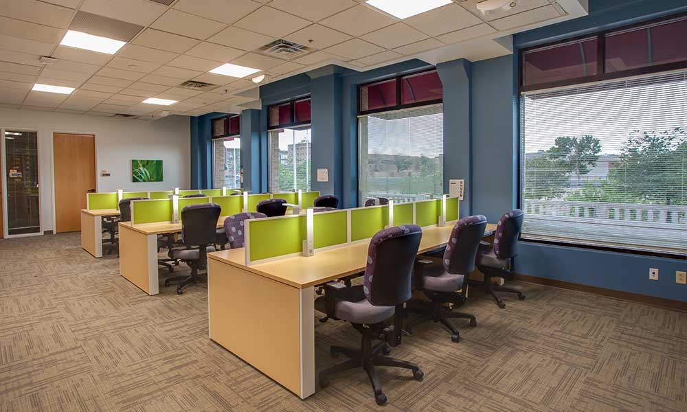 An office space with several workstations featuring green dividers and black office chairs. The room has large windows with blinds, blue accent walls, and a beige carpet. The ceiling has recessed lighting and visible ventilation ducts.