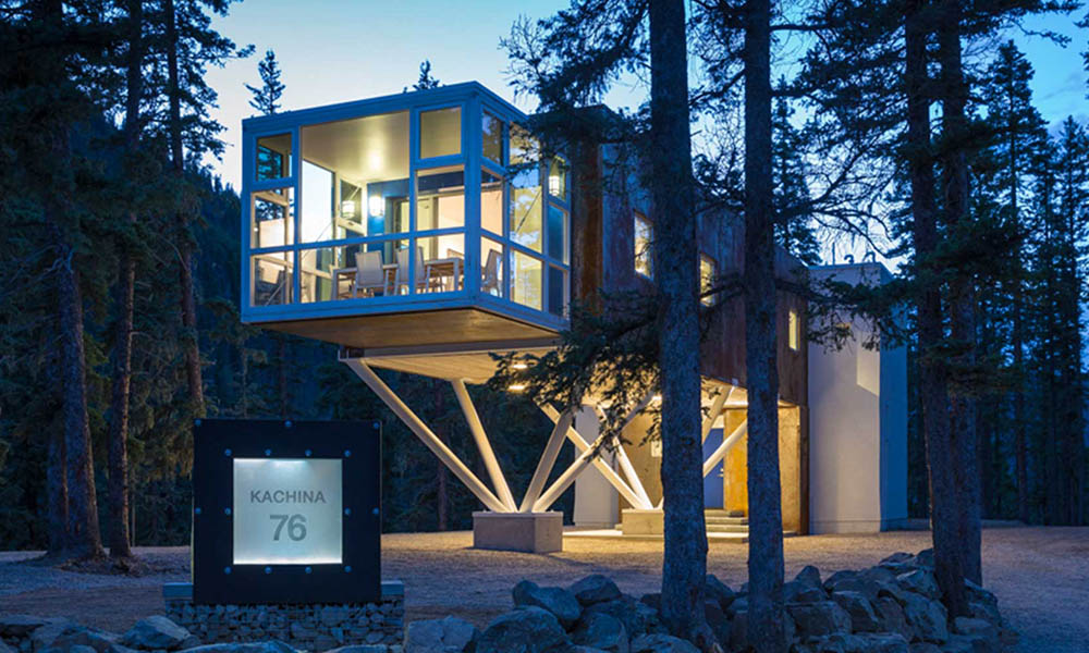 A modern house elevated on stilts is surrounded by tall trees. Large windows on one side reveal the interior, illuminated against the evening sky. A sign at the base reads "Kachina 76." The structure rests on a solid foundation with rock landscaping below.