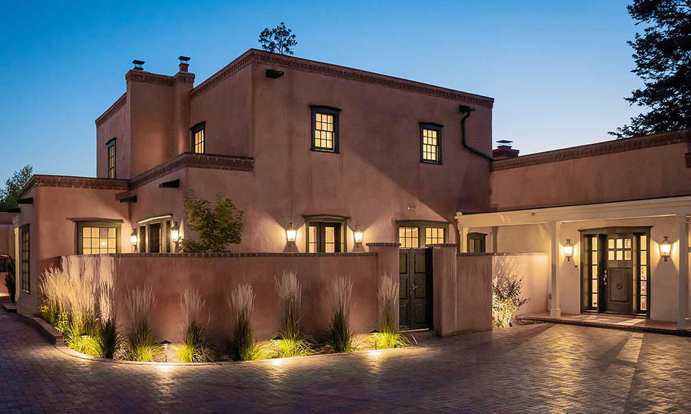 A two-story adobe-style house is shown at dusk with lights illuminating the exterior. The house features flat roofs, small windows, and a courtyard with landscaped plants. The surroundings are calm and serene, with the sky in the background transitioning to night.