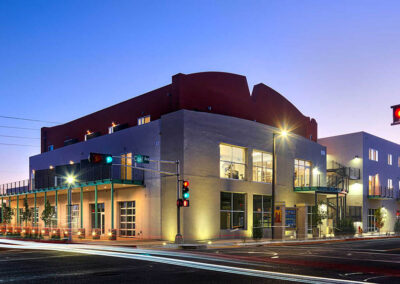 A modern three-story building is shown at dusk with its exterior lights on. The building is situated at a street intersection with a visible traffic light. Several windows are lit up, and there appears to be light traffic on the street.