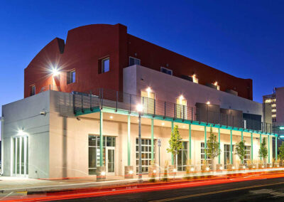 A modern, two-story building with a mix of white and red exterior walls is depicted at dusk. The structure features large windows and a balcony with teal railings. The surrounding area includes streetlights, trees, and a backdrop of additional buildings.